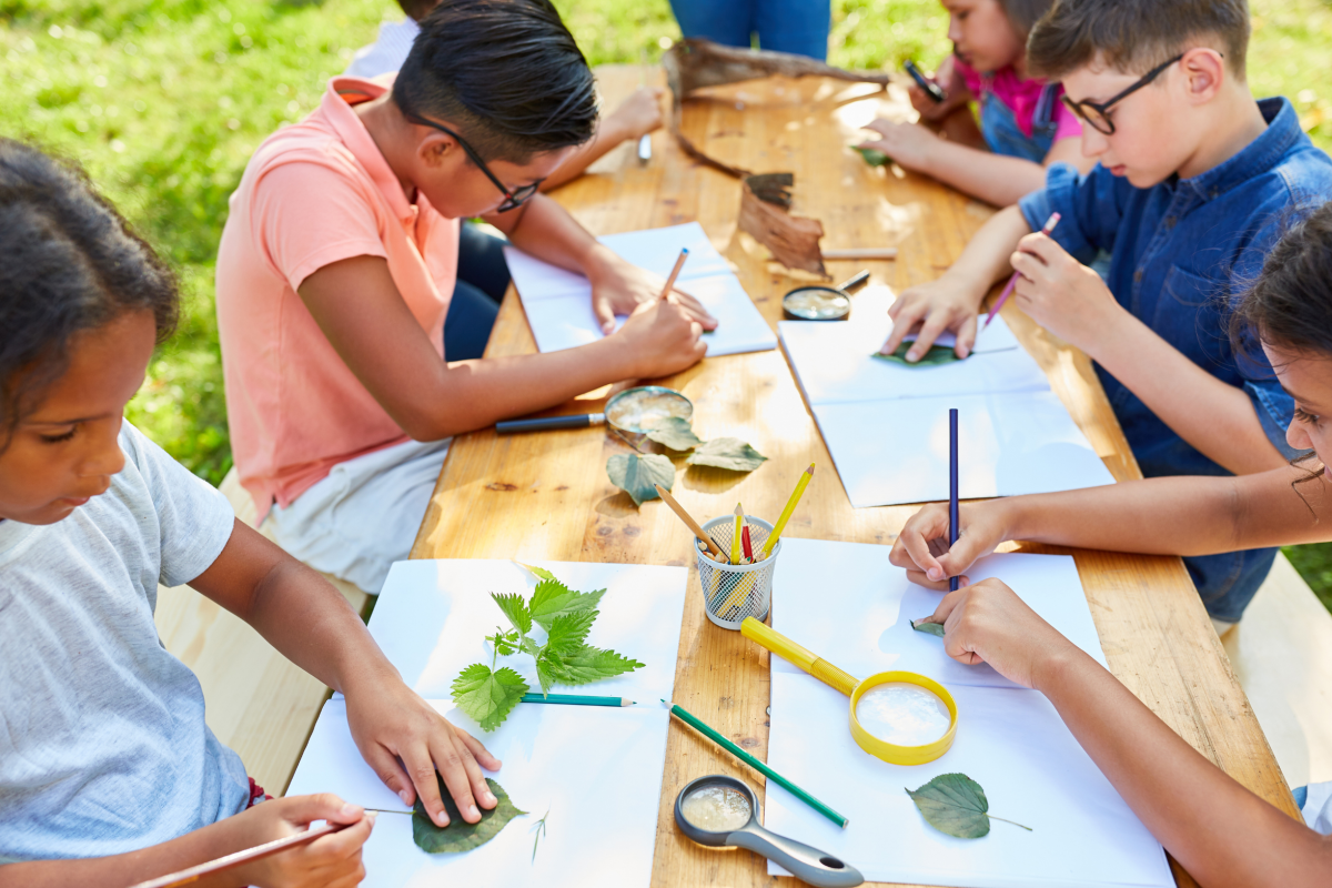 School age children sitting at table outdoors tracing leaves